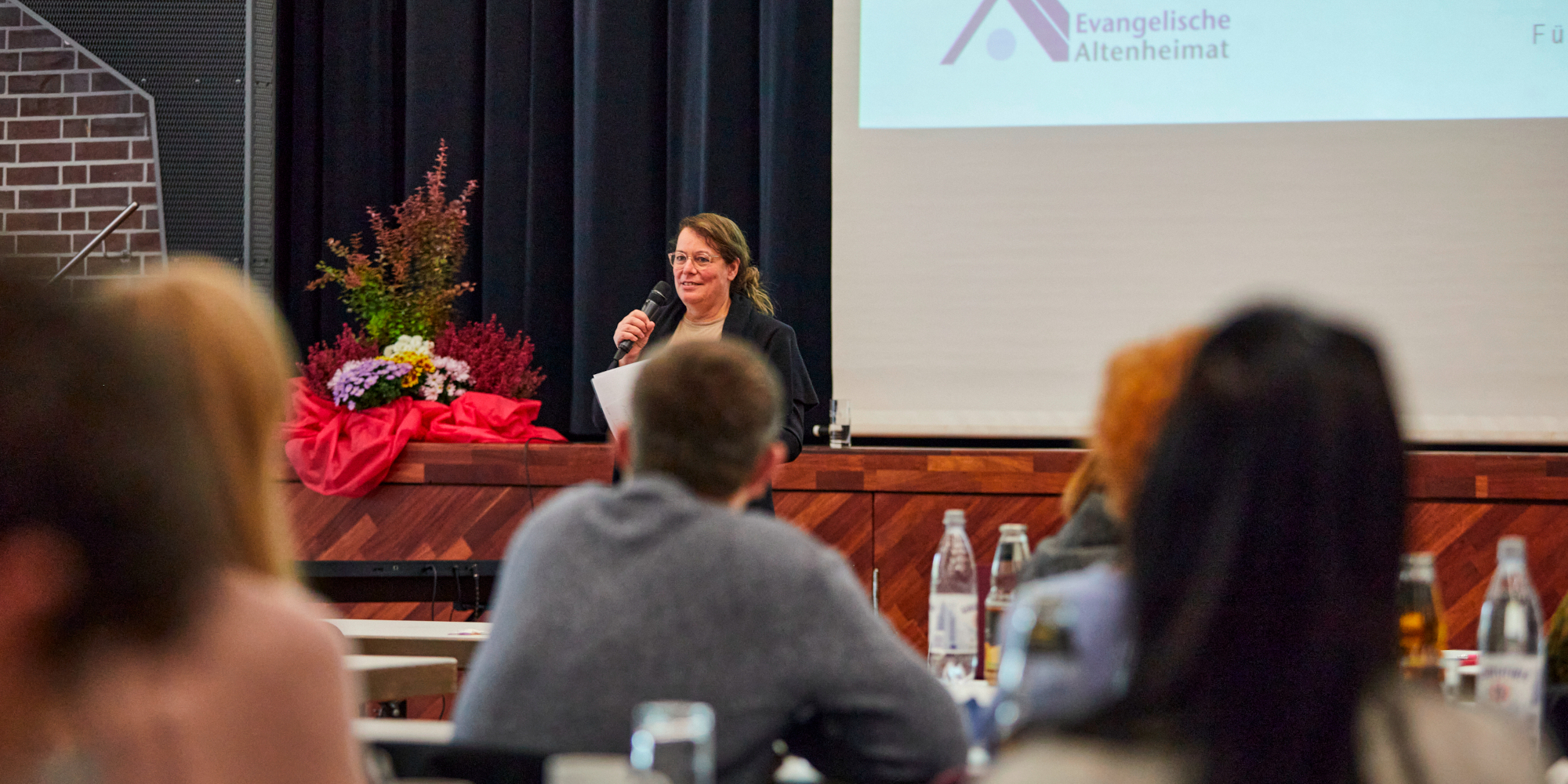 Referntin mit Mikro im Hintergrund vor Beamerpräsentation, fotografiert aus dem Auditorium mit einigen sitzenden Personen von hinten