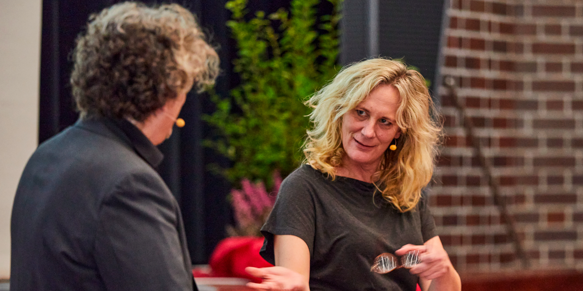 Mann und Frau vor Blumendeko im Theaterspiel, sie mit Brille in rechter Hand mit Gesicht zur Kamera, er von hinten