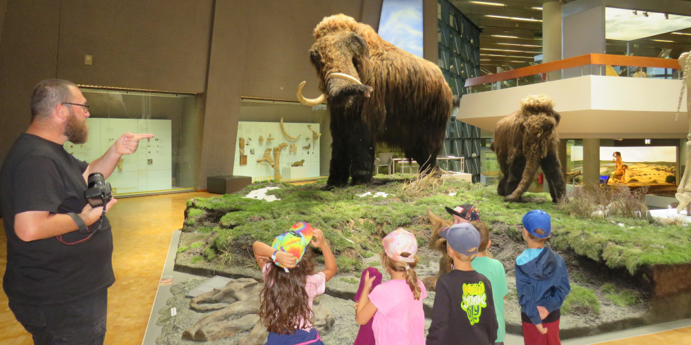 Kinder betrachten ein Mammut im Museum, links unten eine erwachsene Begleitperson.