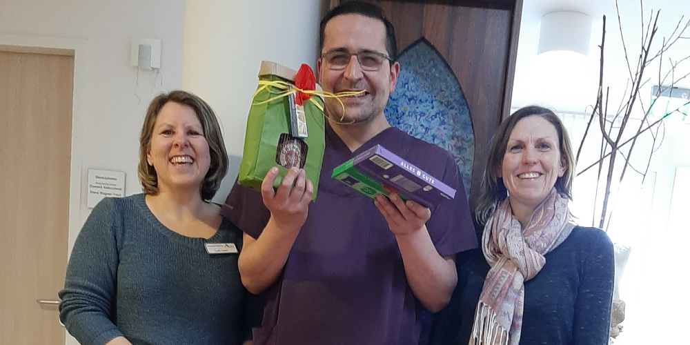 Zwei lachende Frauen mit Mann in Arbeitskleidung in ihrer Mitte, der strahlend Geschenke in den Händen hochhält