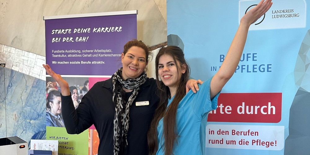 Zwei junge Vertreterinnen der Evangelischen Altenheimat, eine davon in Berufskleidung, vor Werbebannern winkend