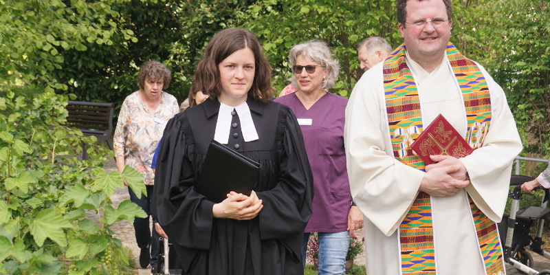 Evangelische junge Pfarrerin im scharzen Talar und katholischer Pfarrer im bunten Ordinat beim Einzug mit Mitarbeitenden