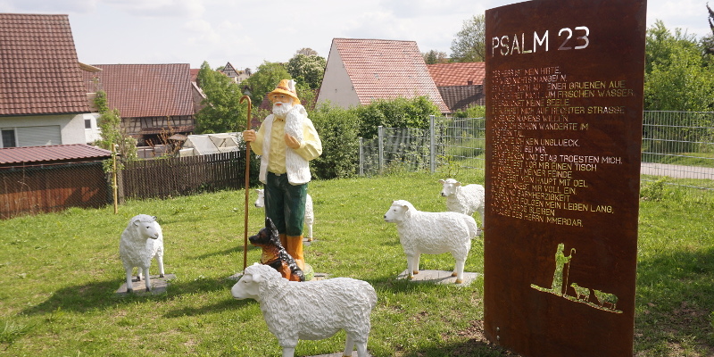 Figurengruppe Hirte mit Schafen und Stele aus rostigem Stahl mit Psalm 23 auf grüner Wiese