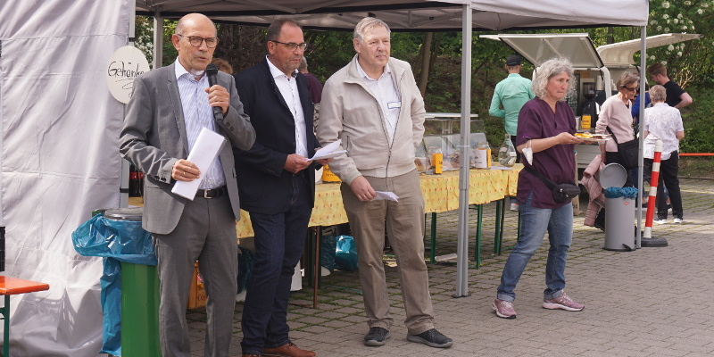 Drei Redner vor Kuchenstand, der linke mit Mikro in der Hand